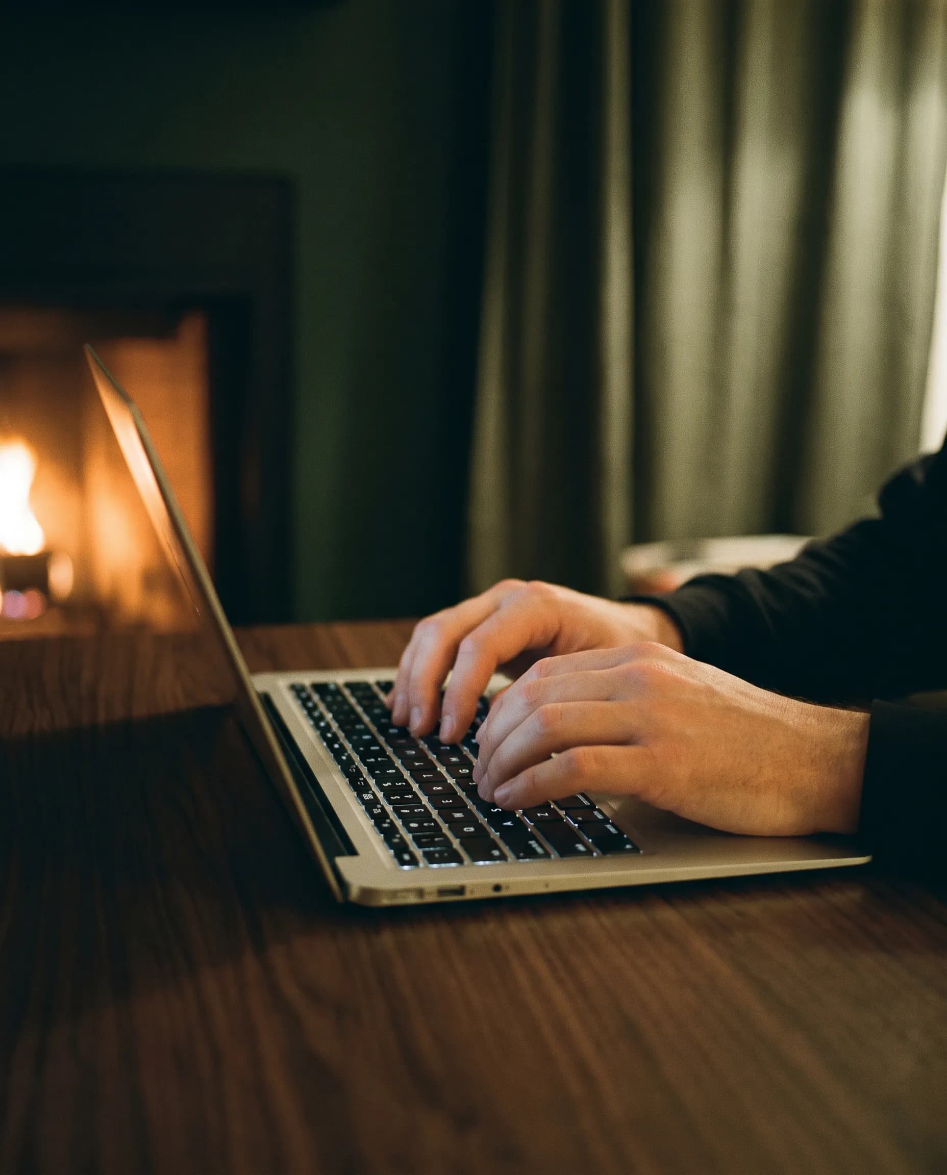 Hands typing on a laptop at a wooden desk beside a fireplace